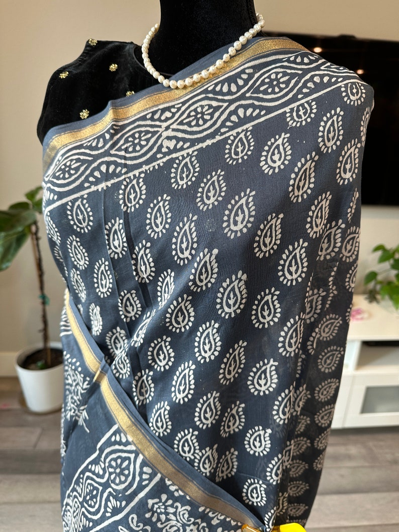 May include: A grey cotton saree with a white paisley print and a gold border. The saree is draped over a mannequin.