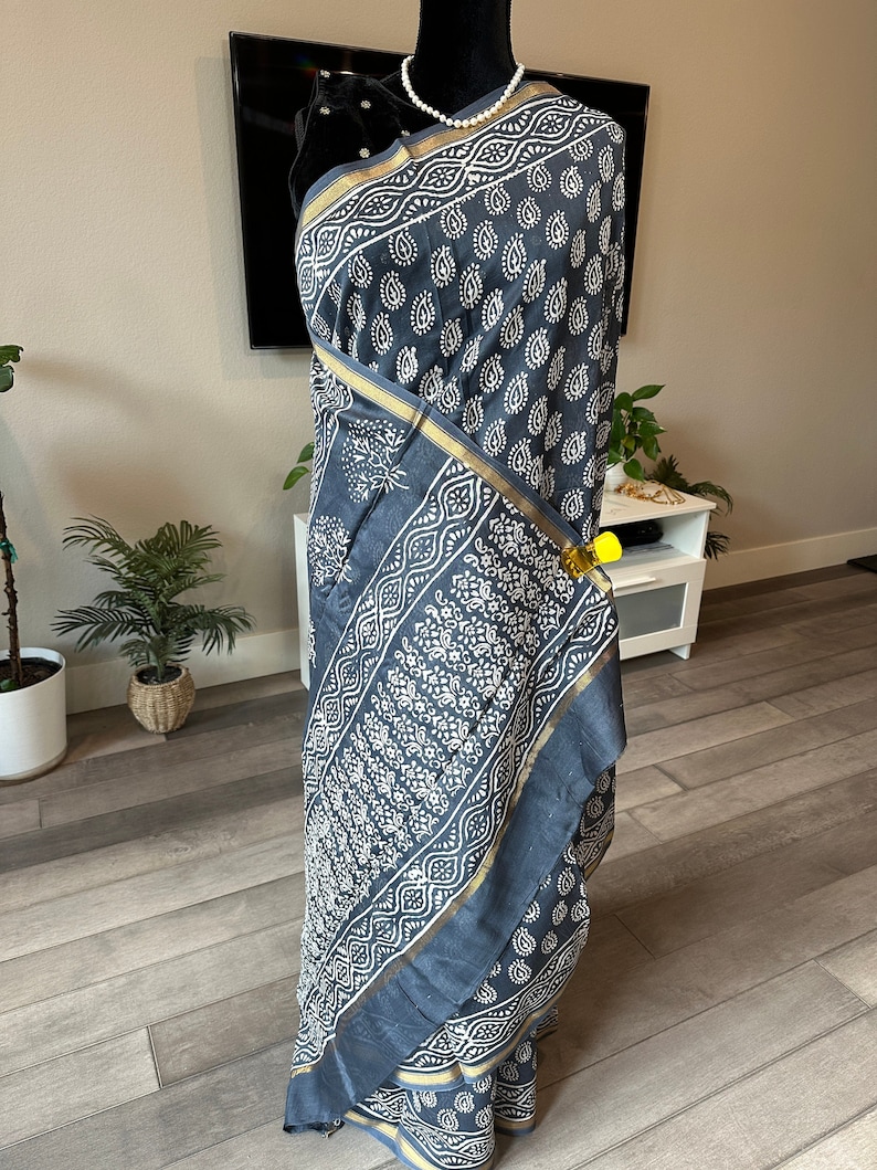 May include: A grey and white cotton sari with a paisley print and gold trim. The sari is draped on a mannequin.