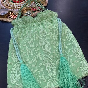 Puede incluir: Una bolsa de cordón de color verde claro con un patrón de paisley y dos borlas largas. La bolsa está hecha de una tela suave y delicada y tiene una parte superior fruncida.