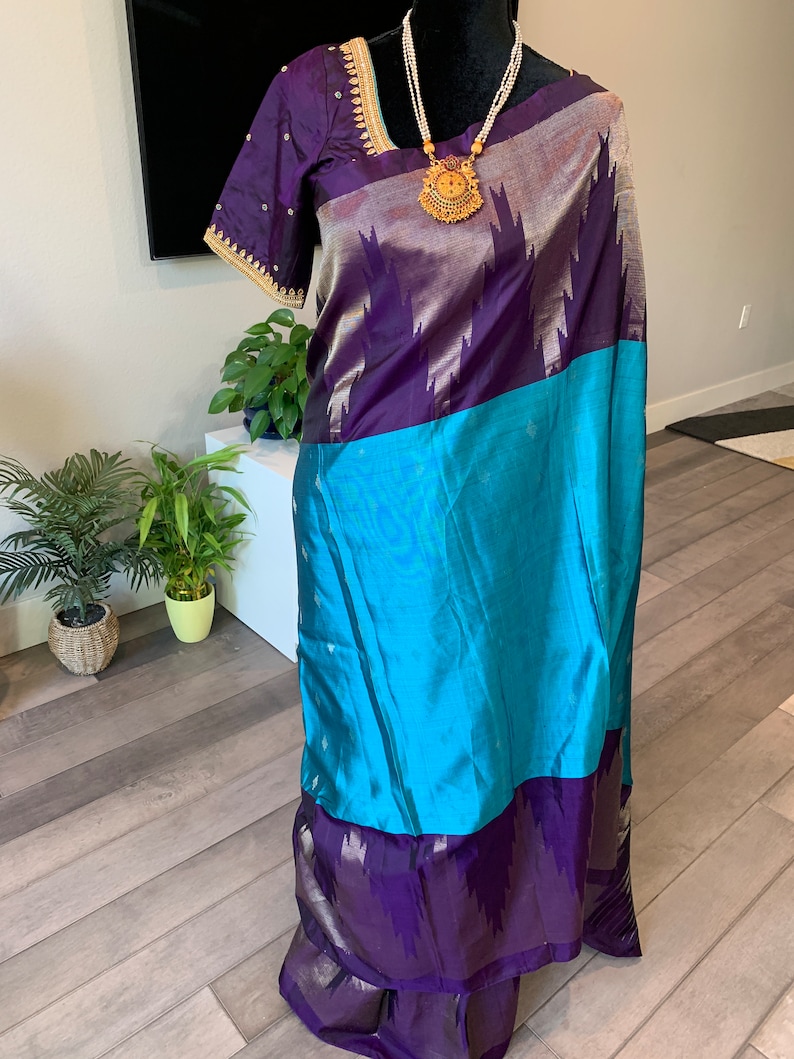 May include: A purple and blue silk saree with gold embroidery and a gold necklace. The saree is draped on a mannequin.