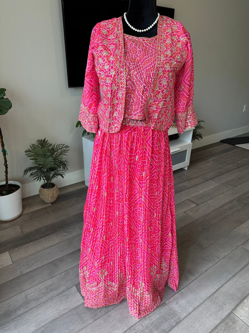 May include: A pink and gold three-piece Indian outfit with a long skirt, a short-sleeved top, and a matching jacket. The outfit is decorated with intricate patterns and sequins.