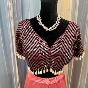 May include: A maroon sequined crop top with a white shell trim and a matching pink skirt. The top has a V-neckline and short sleeves. The skirt is a simple, flowing style. The model is wearing a pearl necklace.