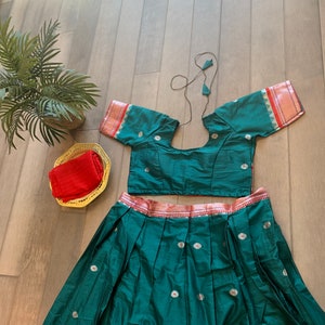 May include: A green skirt and matching crop top with a red and gold trim. The skirt has a pleated design and is decorated with small white flowers. The top has a round neckline and short sleeves.