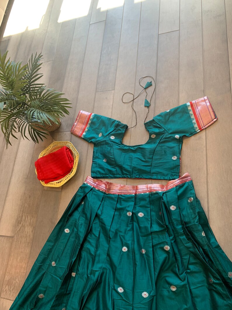 May include: A green skirt and matching crop top with a red and gold trim. The skirt has a pleated design and is decorated with small white flowers. The top has a round neckline and short sleeves.