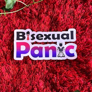 May include: A sticker with the text "Bisexual Panic" in black and white, with the word "Panic" in a gradient of pink, purple, and blue. A small illustration of a person in a maid outfit is on the right. The sticker is on a red, textured surface.