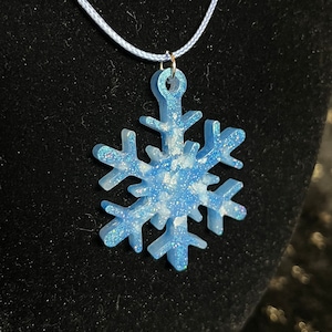 May include: A light blue snowflake pendant necklace with glitter accents. The snowflake is suspended from a light blue cord. The pendant is a winter-themed accessory.