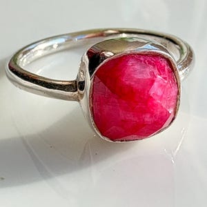 Puede incluir: Un anillo de plata con una gran gema roja brillante facetada. La gema, de forma cuadrada con esquinas redondeadas, está engastada en una sencilla banda de plata pulida. El anillo se muestra sobre una superficie blanca, resaltando el vibrante color de la piedra.