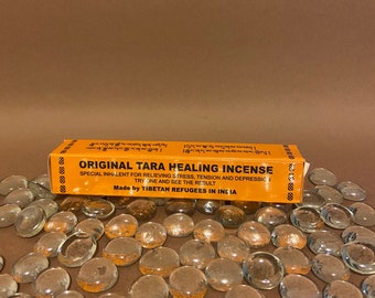 May include: An orange box of "ORIGINAL TARA HEALING INCENSE" with text, surrounded by clear glass pebbles. The box states it is an inhalant for stress, tension, and depression, made by Tibetan refugees in India.
