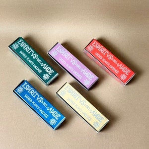 Puede incluir: Cinco cajas rectangulares de incienso en varios colores: verde, rosa, rojo, azul y amarillo. Cada caja tiene el texto "ESPÍRITVS DEL AUDE SACRED PLANTS INCENSE" en una fuente estilizada. Las cajas están dispuestas sobre una superficie de tono neutro.