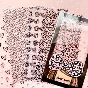 Könnte beinhalten: Vier Blätter aus transparentem Vinyl mit rosa und schwarzen Mustern. Die Muster umfassen Herzen, Schleifen und Leopardenmuster. Die Blätter sind auf einem rosa Hintergrund mit einem goldenen Schleifenmuster angeordnet.