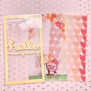 Puede incluir: Un marco dorado con un inserto de plástico transparente que presenta el texto "hello spring" y un cerdo de dibujos animados sosteniendo un paraguas. El marco está sobre un fondo rosa con un patrón repetitivo de lazos dorados.