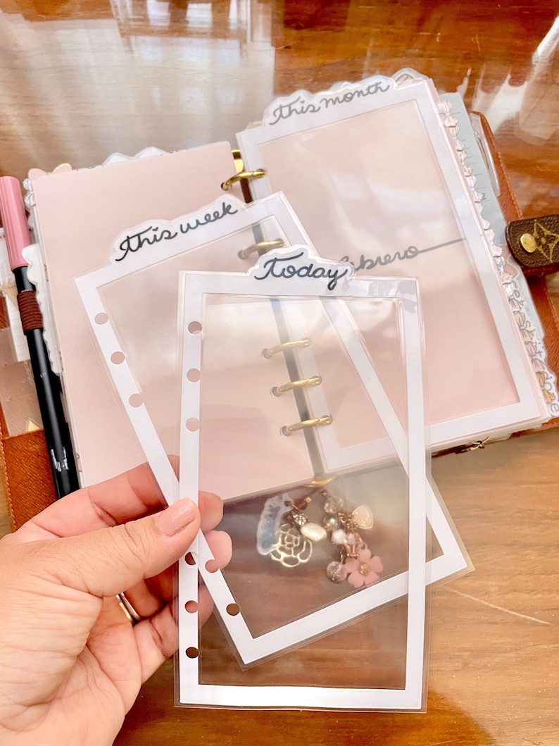 Peut inclure: Trois pochettes en plastique transparent avec des bordures blanches pour un agenda ou un journal. Les pochettes sont &eacute;tiquet&eacute;es "Cette semaine", "Aujourd'hui" et "Ce mois-ci".