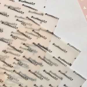 May include: Multiple sheets of translucent paper are printed with the hashtag and word "coffeeaddict" in a repeating pattern. The text is in a simple, sans-serif font and is printed in black ink. The paper has a slight texture.