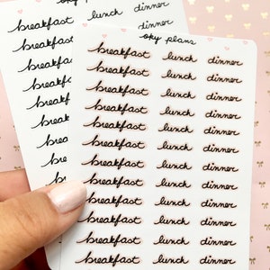 Może przedstawiać: Dwa arkusze przezroczystych naklejek z czarnym ręcznie pisanym tekstem. Naklejki są do planowania posiłków i zawierają słowa "śniadanie", "obiad" i "kolacja".