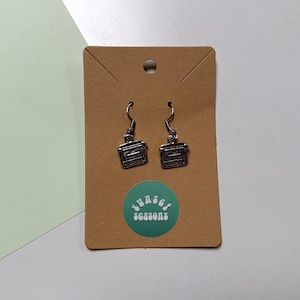 May include: Silver-coloured earrings in the shape of vintage radios. The earrings are suspended from silver-coloured hooks and presented on a brown card with a green circular sticker reading "sunset seasons". The background is light green and white.