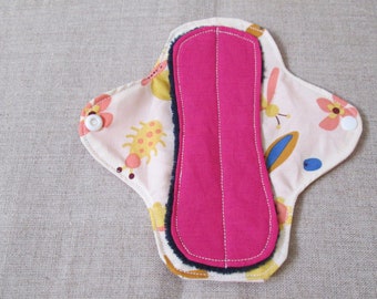 May include: A pink reusable cloth menstrual pad with a floral print and white snaps. The pad is folded in half and has a dark blue and black layer on the outside.