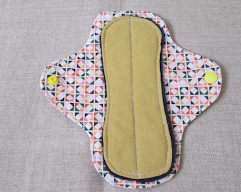 May include: A reusable cloth menstrual pad with a geometric print in shades of pink, red, green, and black. The pad has a yellow snap on each side and a brown absorbent layer.