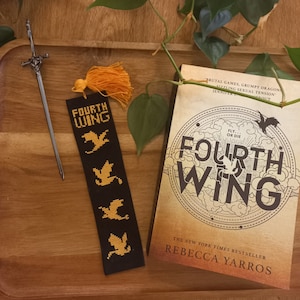 May include: A black bookmark with a gold tassel and embroidered dragons. The bookmark features the text "Fourth Wing" and the words "Fly... or Die".