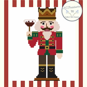 May include: A cross-stitch pattern of a nutcracker wearing a gold crown, red jacket with green accents, and black boots, holding a stemmed glass. The design features a circular logo in the top right corner with the text AJ Threadworks Since 2026.