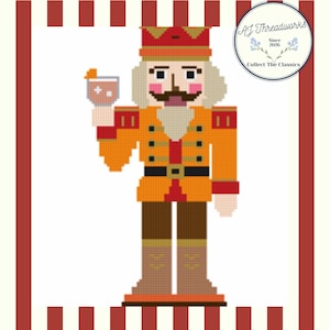 May include: A pixel art cross-stitch pattern of a nutcracker wearing an orange uniform with a black belt, holding a cocktail glass with an orange slice. The design features a circular logo in the top right corner with the text AJ Threadworks Since 2026 Collect The Classics.