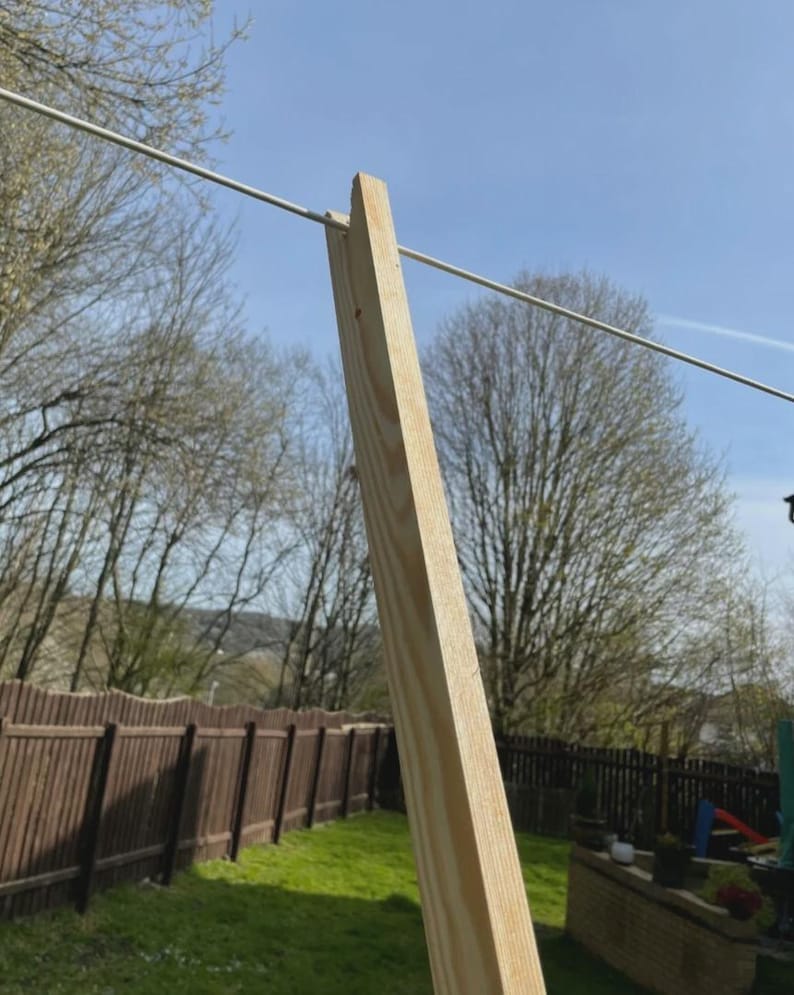 K&ouml;nnte beinhalten: Ein Holzpfosten mit einem daran befestigten Metalldraht, wahrscheinlich f&uuml;r eine W&auml;scheleine. Der helle Holzpfosten steht vor einem Hintergrund aus gr&uuml;nem Rasen, einem braunen Zaun und einem klaren blauen Himmel.