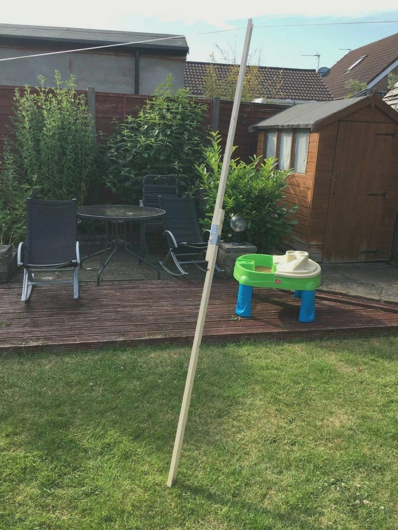 K&ouml;nnte beinhalten: Ein langer, unfertiger Holzpfahl steht in einem Garten. Der Pfahl ist hell und scheint ein Baumaterial zu sein. Ein kleiner, farbenfroher Kinderspieltisch ist ebenfalls zu sehen.