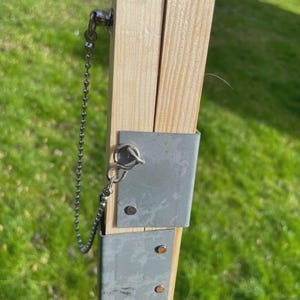 K&ouml;nnte beinhalten: Nahaufnahme einer Holzkonstruktion mit Metallhalterungen und einer Kette. Das Holz ist hell und das Metall ist silberfarben. Die Kette ist mit einem kleinen Ring am Holz befestigt. Der Hintergrund ist verschwommenes gr&uuml;nes Gras.