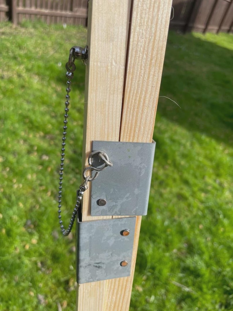 K&ouml;nnte beinhalten: Nahaufnahme einer Holzkonstruktion mit Metallhalterungen und einer Kette. Das Holz ist hell und das Metall ist silberfarben. Die Kette ist mit einem kleinen Ring am Holz befestigt. Der Hintergrund ist verschwommenes gr&uuml;nes Gras.