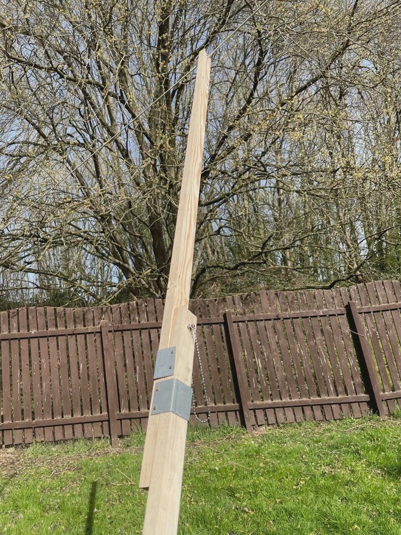 K&ouml;nnte beinhalten: Ein hoher, unfertiger Holzpfosten mit Metallhalterungen und einer Kette, wahrscheinlich f&uuml;r ein Tor oder einen Zaun. Er steht vor einem Hintergrund aus einem braunen Holzzaun, gr&uuml;nem Gras und einem Baum mit Knospen. Das Holz hat eine nat&uuml;rliche, helle Farbe.