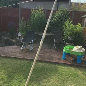 K&ouml;nnte beinhalten: Ein langer, unfertiger Holzpfahl steht aufrecht in einem Garten. Der Pfahl ist hell und scheint Teil einer gr&ouml;&szlig;eren Struktur zu sein. Im Hintergrund befinden sich eine Holzterrasse, Gartenm&ouml;bel und ein kleiner Spieltisch.