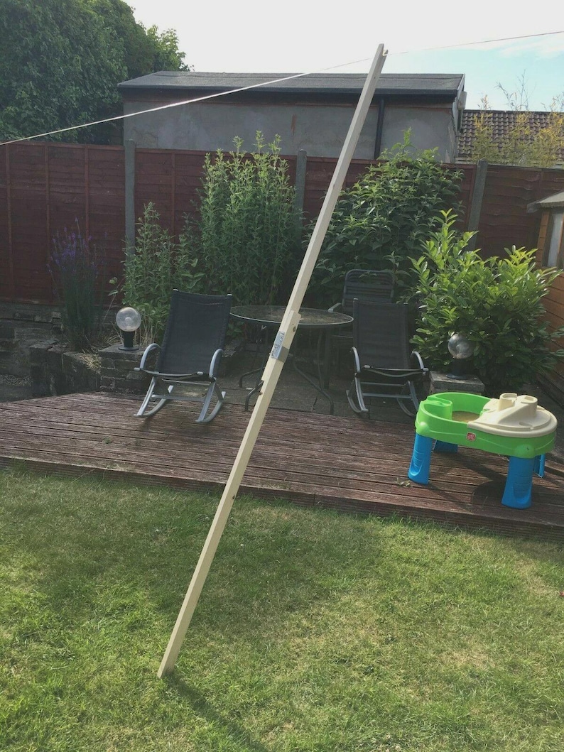 K&ouml;nnte beinhalten: Ein langer, unfertiger Holzpfahl steht aufrecht in einem Garten. Der Pfahl ist hell und scheint Teil einer gr&ouml;&szlig;eren Struktur zu sein. Im Hintergrund befinden sich eine Holzterrasse, Gartenm&ouml;bel und ein kleiner Spieltisch.