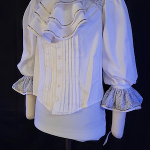 May include: White blouse with a high collar, ruffled front, and long sleeves. The blouse features gold trim on the ruffles and cuffs, with lace detailing on the shoulders and cuffs. A decorative brooch accents the collar.