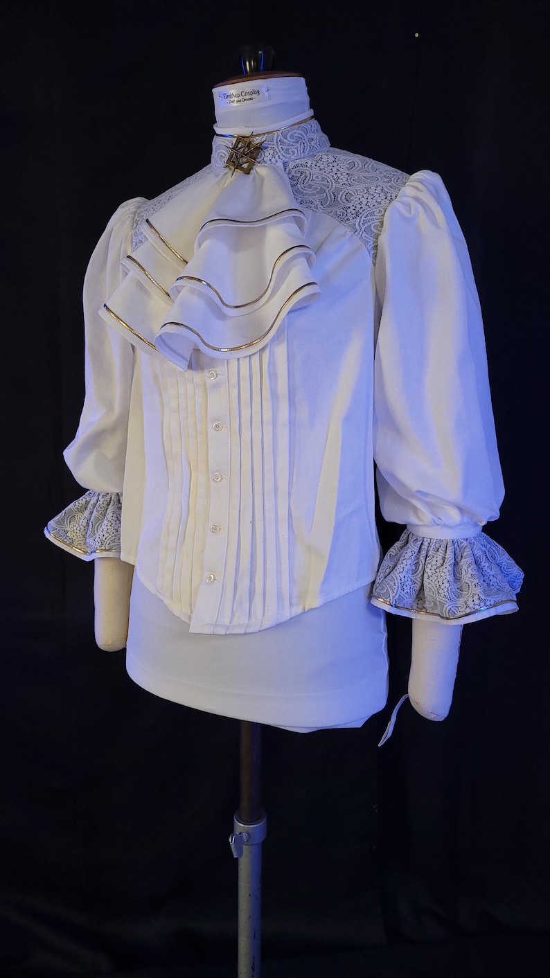 May include: White blouse with a high collar, ruffled front, and long sleeves. The blouse features gold trim on the ruffles and cuffs, with lace detailing on the shoulders and cuffs. A decorative brooch accents the collar.
