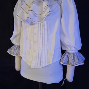 Op de afbeelding: Witte blouse met een hoge kraag, ruches aan de voorkant en lange mouwen. De blouse heeft gouden randen op de ruches en manchetten, met kanten details op de schouders en manchetten. Een decoratieve broche accentueert de kraag.