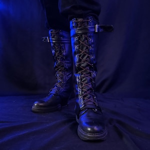 May include: Black leather knee-high boots with brown laces and buckle straps. The boots have a rugged, combat-style design with a thick sole. The image is set against a dark blue background, highlighting the boots' details.