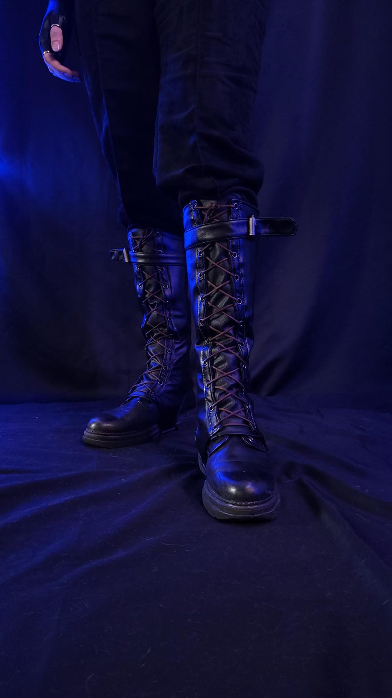 May include: Black leather knee-high boots with brown laces and buckle straps. The boots have a rugged, combat-style design with a thick sole. The image is set against a dark blue background, highlighting the boots' details.