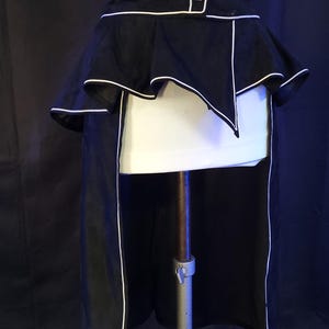 May include: A black and white corset-style garment with a high waist and a peplum design. The corset features white piping along the edges and a button closure. The peplum has a layered, ruffled effect. The garment is displayed on a mannequin.