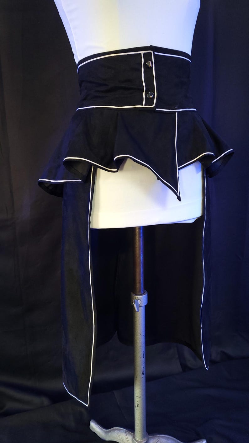 May include: A black and white corset-style garment with a high waist and a peplum design. The corset features white piping along the edges and a button closure. The peplum has a layered, ruffled effect. The garment is displayed on a mannequin.