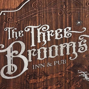 Puede incluir: Cartel de madera marrón con el texto "The Three Broomsticks Inn & Pub" en una fuente decorativa. El cartel tiene un borde blanco y presenta ilustraciones de escobas, el número "3", el texto "Hogsmeade" y el año "1192".