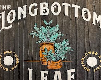 May include: A dark wooden sign with white and gold lettering. It reads "THE LONGBOTTOM LEAF" and features illustrations of plants in a barrel and a basket. The sign also says "PIPE WEED" and "FINEST IN THE SHIRE".