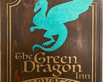 May include: A wooden sign featuring a teal dragon silhouette and gold lettering. The text reads "The Green Dragon Inn, Bywater, The Shire." The sign has a dark brown background and a gold border.
