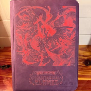 Puede incluir: Un archivador de cartas coleccionables morado con un gráfico rojo de una criatura de fuego. El archivador tiene el texto "MEGA EVOLUTION PHANTASMAL FLAMES GUARD" en rojo. El archivador rectangular es de material similar al cuero.
