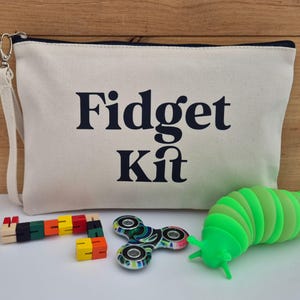 May include: A cream canvas pouch with the words "Fidget Kit" in navy blue. The kit includes a wooden block puzzle, a colourful fidget spinner, and a green caterpillar-shaped fidget toy.