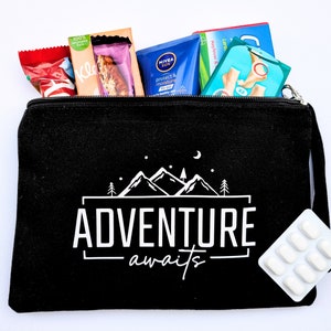 May include: Black canvas zippered pouch with the text "Adventure awaits" in white. The pouch is filled with various items, including a blue sunscreen bottle, a red and white candy bar, a white pill pack, and other items.
