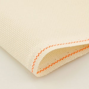 Peut inclure: Gros plan sur une toile Aida blanche avec une bordure cousue orange. Le tissu est tiss&eacute; avec un motif en grille, id&eacute;al pour les projets de point de croix.
