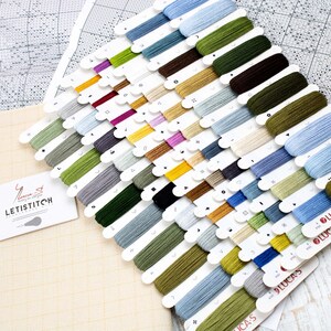 May include: A cross-stitch kit featuring a selection of colourful thread spools. The threads are arranged in rows and include shades of green, blue, brown, and purple. A canvas and pattern are also visible, along with a business card that says "LETISTITCH".