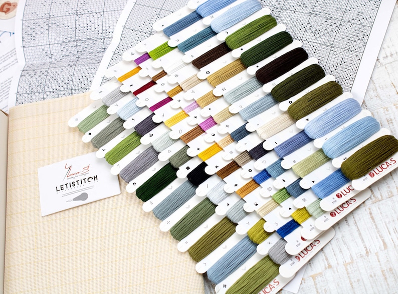 May include: A cross-stitch kit featuring a selection of colourful thread spools. The threads are arranged in rows and include shades of green, blue, brown, and purple. A canvas and pattern are also visible, along with a business card that says "LETISTITCH".