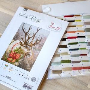 May include: A cross-stitch kit featuring a reindeer with a red bow and holly wreath. The kit includes a white Aida cloth, DMC embroidery floss, and a needle. The kit is labeled "Cerb" and is made in Moldova.