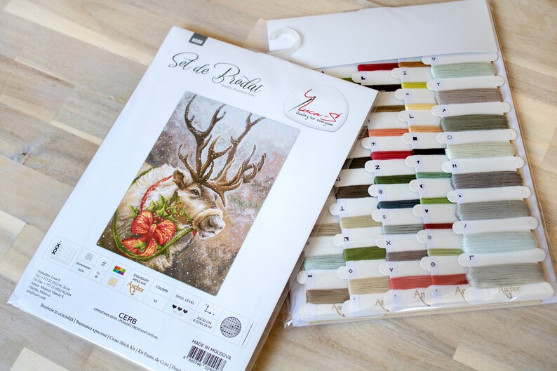 May include: A cross-stitch kit featuring a reindeer with a red bow and holly wreath. The kit includes a white Aida cloth, DMC embroidery floss, and a needle. The kit is labeled "Cerb" and is made in Moldova.