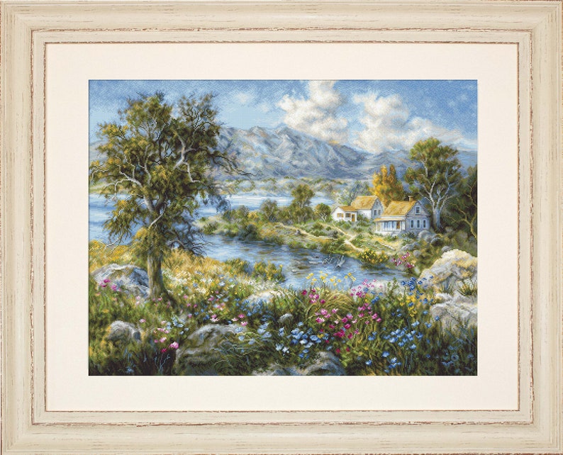 May include: Framed landscape art depicting a tranquil river scene with a tree, houses, and mountains. The artwork has a cream frame and shows a vibrant meadow with colourful flowers and lush greenery. The sky is blue with fluffy clouds, creating a peaceful vista.